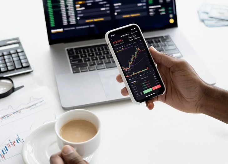 A person studies stock market information on a laptop and mobile phone while enjoying a cup of coffee in a cafe.